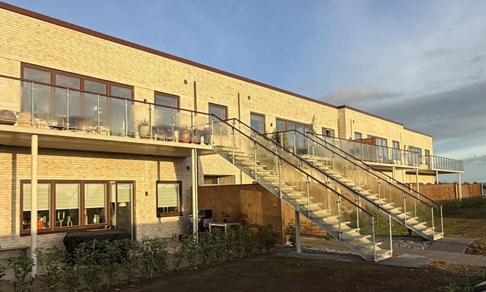 Foto: facade i aftensol med beboede altaner, glasværn og to udvendige trapper til første sal